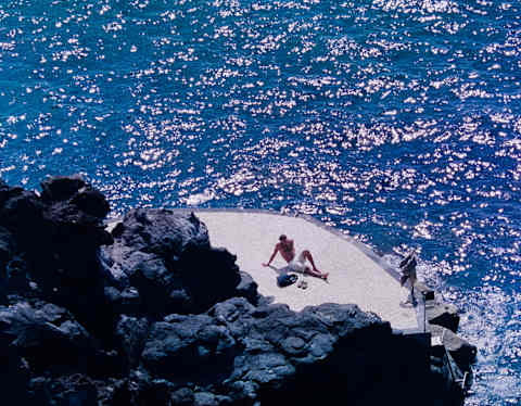 Two people relax on a small, circular concrete platform by the edge of a rocky coastline, surrounded by sparkling blue ocean water under bright sunlight.