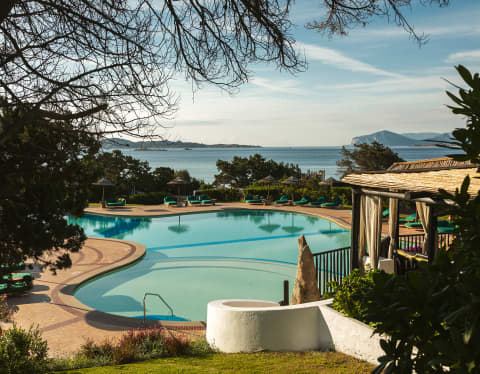 Looking down from green sloping gardens to the tropical pool below, with views past the sun-loungers and parasols to the sea.