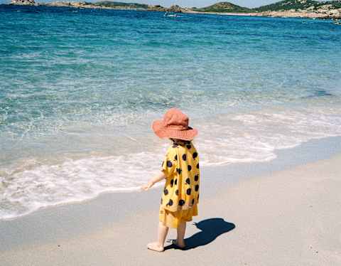 A small child in a yellow and black spotty top and red sunhat approaches the white foam shallows of the bright turquoise sea.