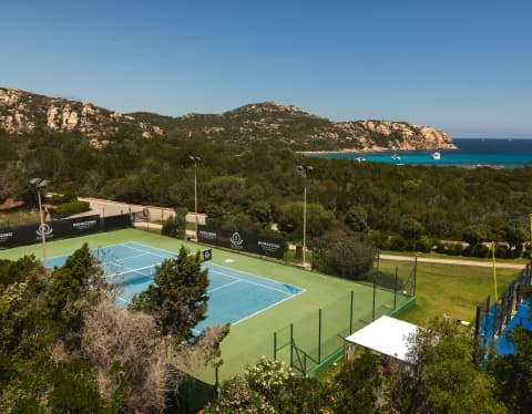 A rocky headland and flashes of bright azure ocean are seen behind a coastal forest, in a view from above the tennis court.