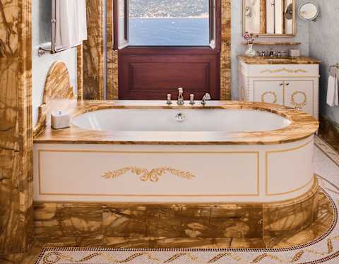A luxurious bathroom with a gold-trimmed bath, ornate marble details, a chandelier, a large mirror, and a window offering a scenic view of the sea and distant hills.