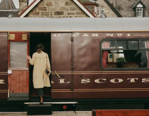 Viewed from behind, a woman in a cream coat uses a portable step to board the Royal Scotsman at Boat of Garten station.