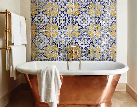 Blue and yellow geometric patterned tiles fill one wall. A free standing copper and ceramic bath reflects the marble floor