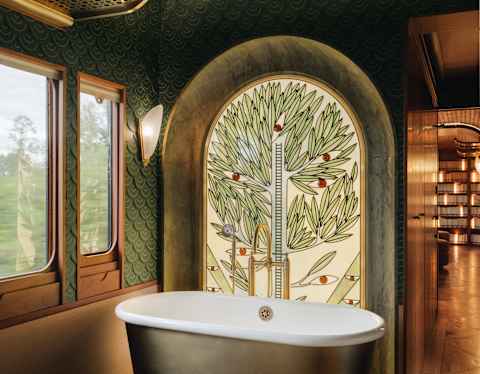 A stained glass depiction of a tree, by Franz Mayer of Munich, rises behind a brass freestanding bathtub with white enamel.