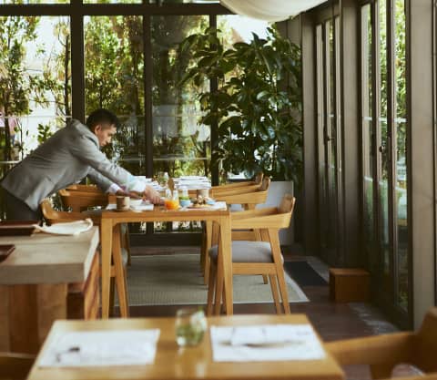 A waiter serves breakfast in a view of a row of wooden tables in Muaka's light-filled dining room with shade canopies.