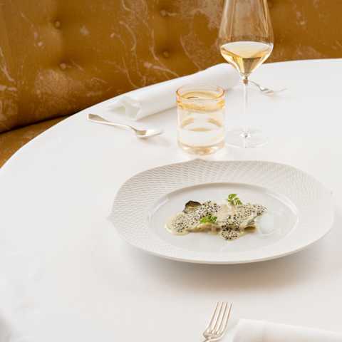 A white tablecloth with a plated gourmet dish, wine glasses, water glasses, and cutlery, set on a round table with a brown upholstered background and a small flower arrangement.