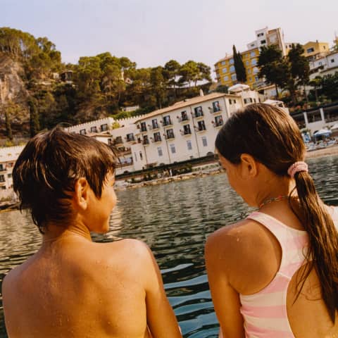 Angled image of a young girl and boy, wet from swimming, sitting in sun on a boat facing Mazzaro beach, viewed from behind.