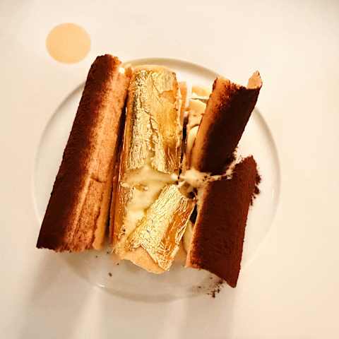 A close-up of a plated dessert featuring layers of sponge and cream, topped with gold leaf, on a white plate with a decorative sauce dot.
