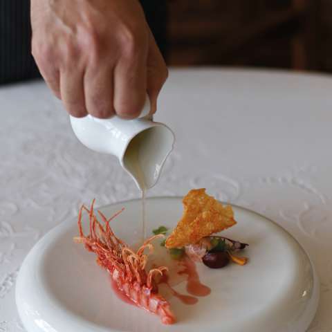 A waiter pours grapefruit tea on to a prawn with a savoury crisp, served on a chunky, shallow white bowl, seen in close-up.