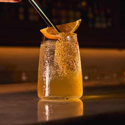 A hand places a straw into a tall glass filled with a sparkling drink and ice, garnished with an orange slice, on a dimly lit bar counter.