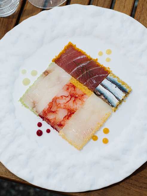 A white plate holds a square mosaic of colourful raw seafood slices—tuna, prawn, white fish, and sardine—with small drops of yellow and red sauces, set on a wooden table.