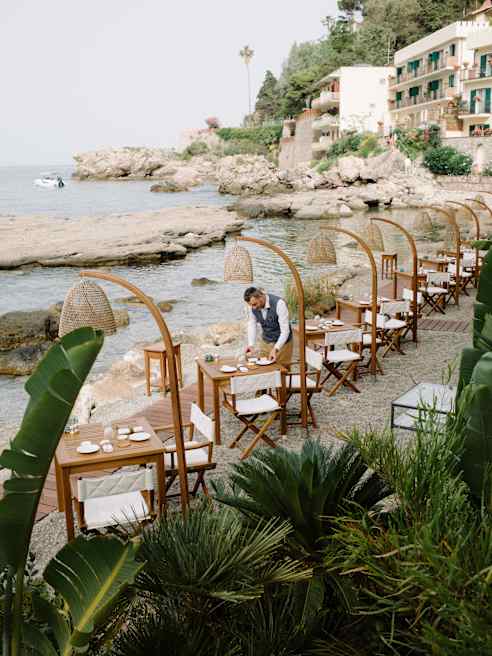 Outdoor dining area with wooden tables and chairs lined up along a rocky shoreline, each table shaded by a woven canopy. Lush green plants in the foreground, and a coastal village and sea in the background.