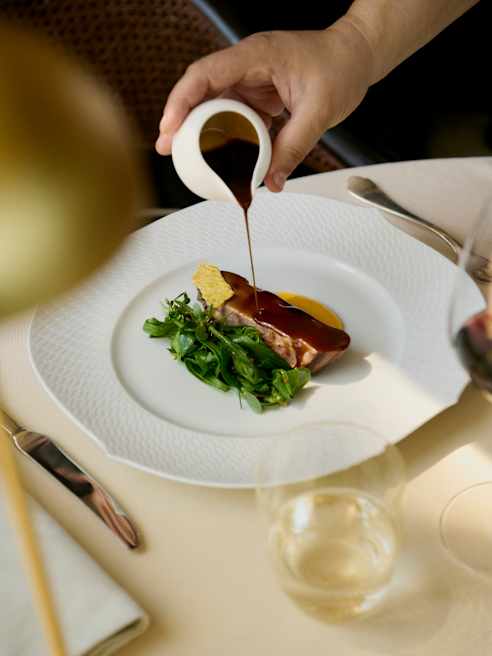 A waiter drizzles rich gravy from a small jug onto a dish of grilled lamb loin with steamed wild chicory greens at Oro.