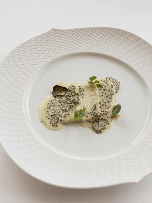 A white textured plate holds a small portion of creamy sauce topped with black caviar, garnished with green herbs and a bay leaf, on a plain white background.