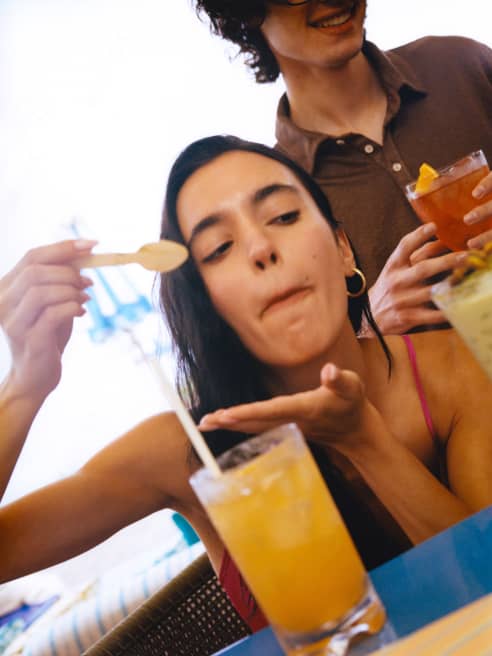 A dark-haired girl raises a hand to catch drips as she tastes an orange drink with a spoon and a man behind holds a Negroni.