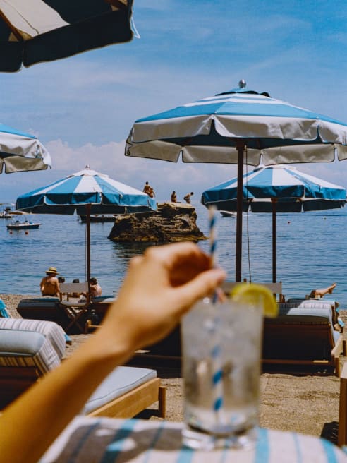 A guest stirs a lemon-garnished drink in soft-focus foreground as swimmers gather on a rock out to sea, beyond the parasols.