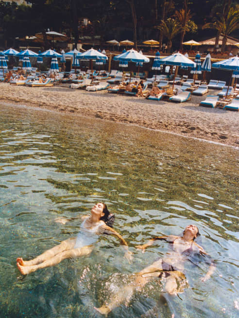 In front of the beachfront parasols, two women in swimming costumes sit in the shallows, heads tipped back, seen from above.