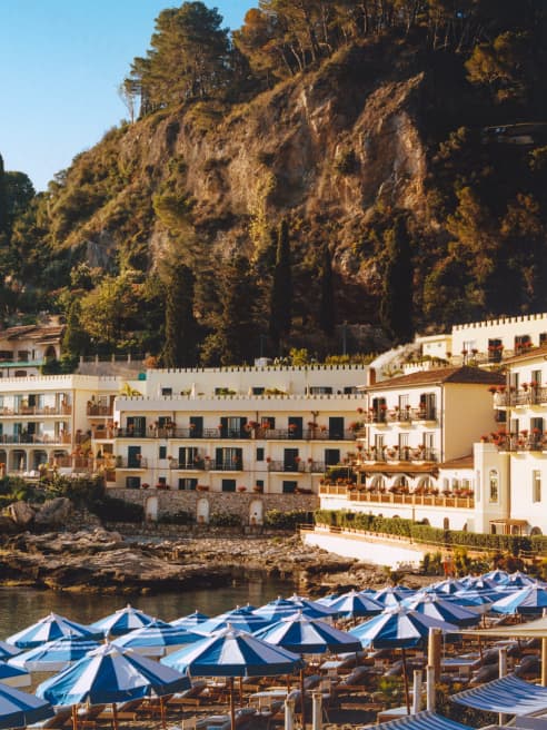 View over blue striped parasols of Lido Villeggiatura to Villa Sant'Andrea hotel, nestled between ocean and forested cliffs.