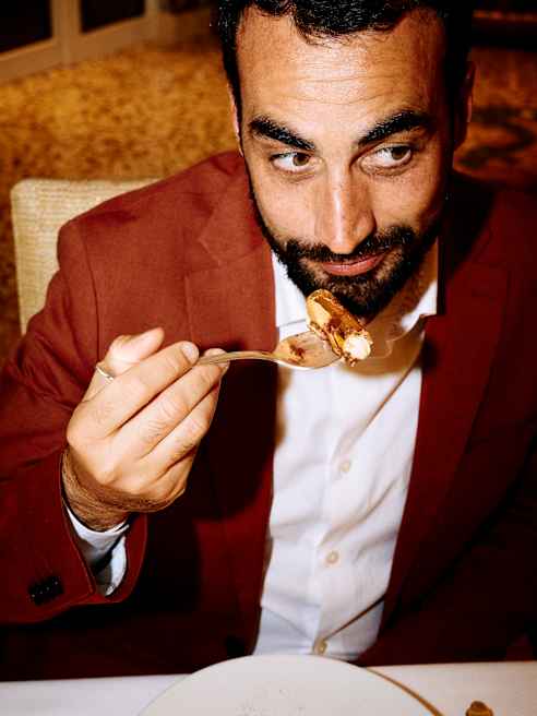 A man in a red blazer sits at a table, holding a fork with pudding. He looks at the food with a slight smile. A partially eaten pudding is on a white plate in front of him.