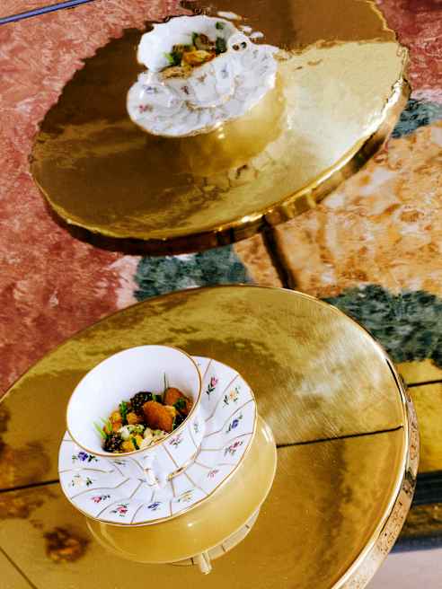 Two ornate white bowls with food are placed on gold, circular tables. The bowls have floral patterns and contain colourful, artfully arranged dishes. The background includes reddish and greenish marble textures.