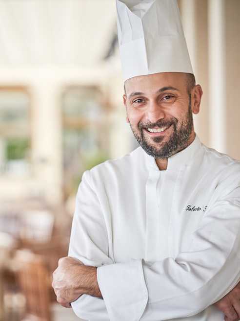 Chef Roberto Toro, with dark facial hair and wearing monogrammed whites and a toque, smiles to camera with his arms crossed.