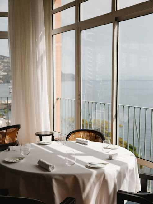 A round window table with four chairs, water glasses and rolled napkins catches the sun and sea views at William Restaurant.