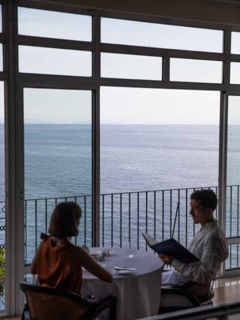 Two people sit at a table by large windows in a restaurant, overlooking a calm sea. One person appears to be reading a menu while the other listens. The scene is peaceful and sunlit.