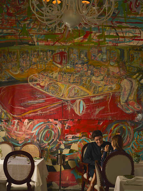 Two people sit at a table in a dimly lit restaurant with ornate chairs, beneath a colourful, abstract mural depicting a red car and various faces. A chandelier hangs from the ceiling above them.