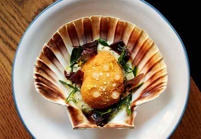 A golden, breaded scallop served in a half shell on a white plate, garnished with salad leaves, set on a wooden table.