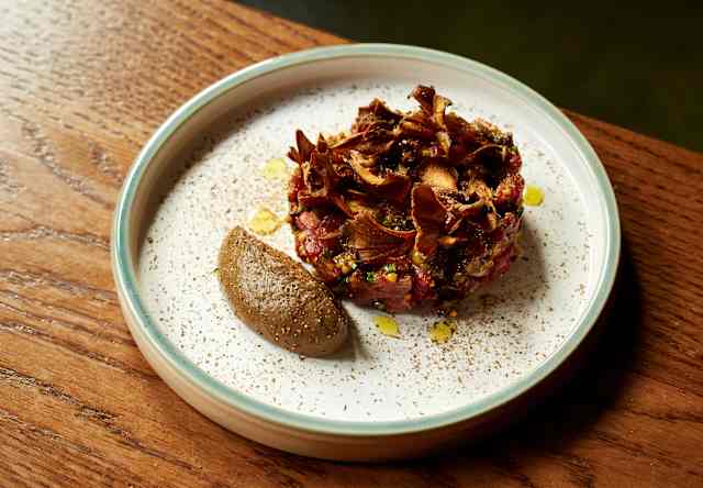 A white plate with a round serving of steak tartare topped with crispy mushrooms, garnished with herbs and oil drops, and a quenelle of dark brown mousse on the side, set on a wooden table.