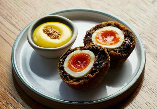 A plate with two halved Scotch eggs, showing soft yolks, next to a small pot of yellow dipping sauce with seeds, set on a wooden table.
