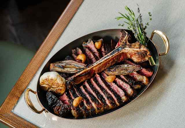 Sliced T-bone steak served on an oval metal platter with roasted garlic and herbs.