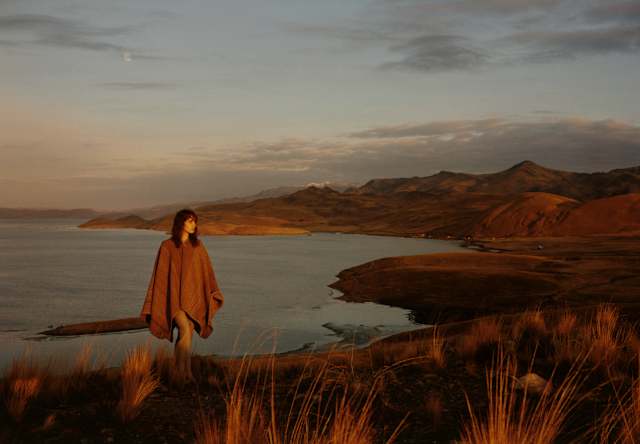 A woman in a brown wool poncho strides up an incline with a backdrop of hill chains and calm lake waters, seen at sunset.