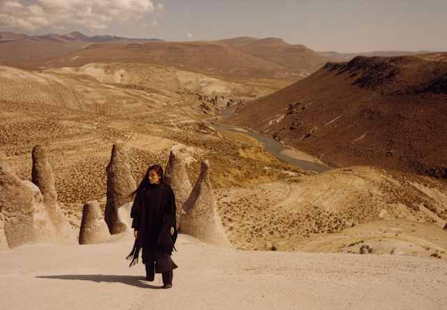 A woman with long dark hair and black clothes stands on a hill overlooking the Puruña stone forest and a valley river beyond.
