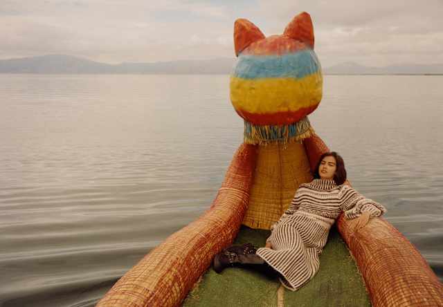 A dark-haired woman in a striped dress lies back on a Lake Titicaca boat made of reeds with a large cat-like figurehead.