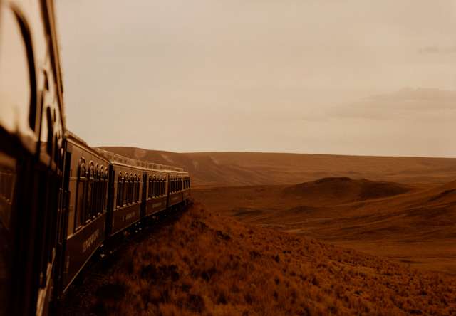External view from the fifth carriage window capturing the train front forging through grasslands tinted deep-red by sunset.