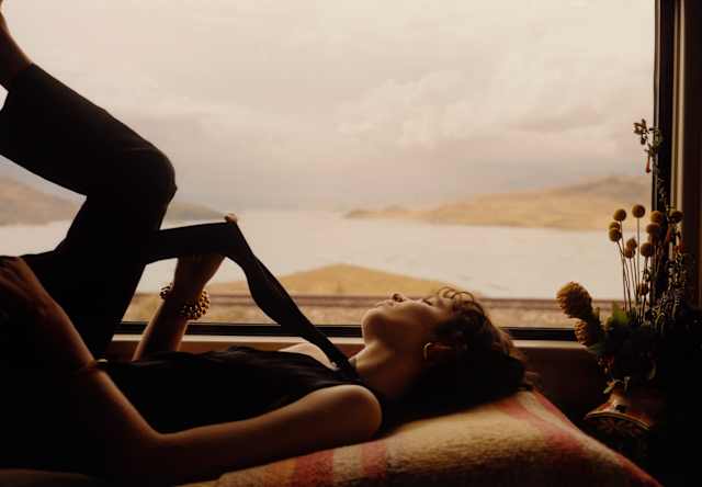 A woman reclines on bed by the window, holding a sash from her top as soft sun plays on her face and lake contours behind.