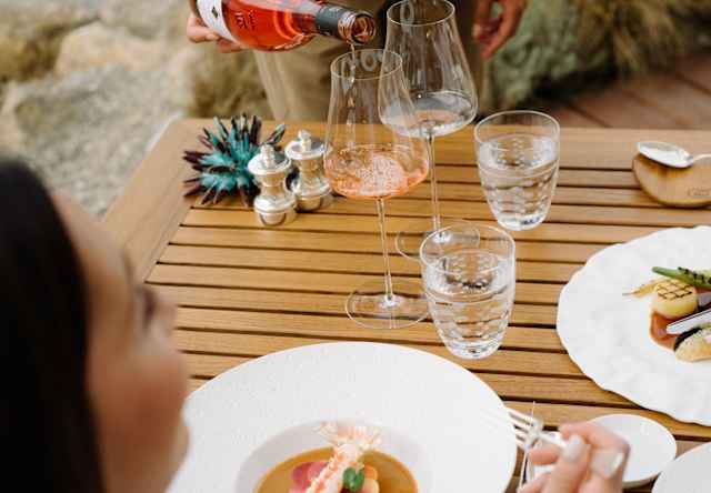 A person seated at an outdoor wooden table is about to eat soup, while another person pours rosé wine into a glass. Plates of gourmet food and glasses are arranged on the table.