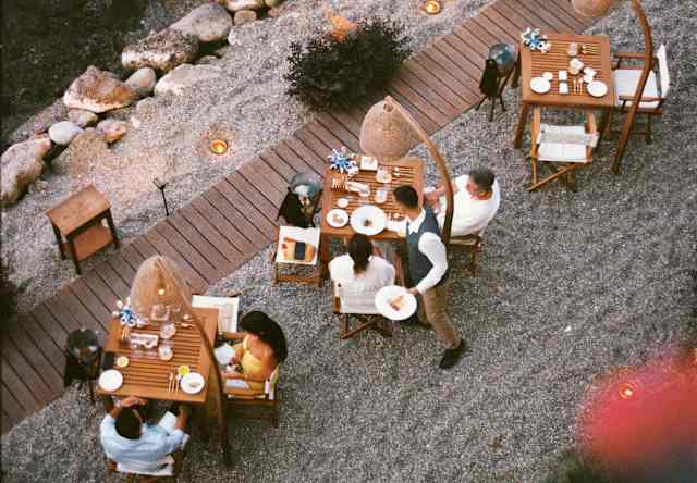 Aerial view of people dining at outdoor wooden tables on a gravel patio by a rocky shoreline, with warm lighting and greenery surrounding the area.