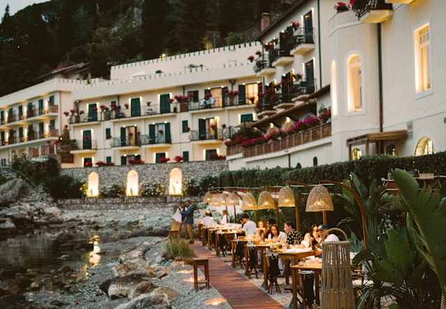 Elegant seaside restaurant set along a rocky shore at dusk, with tables lined up on a wooden walkway, glowing lanterns, and white buildings with balconies rising against a cliff in the background.