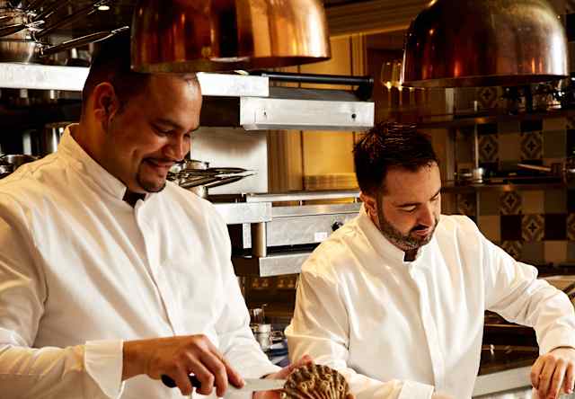 Two chefs in white uniforms prepare food together in a professional kitchen, one slicing a large mushroom and the other arranging ingredients on a tray. Stainless steel cookware and utensils are visible on the worktops.