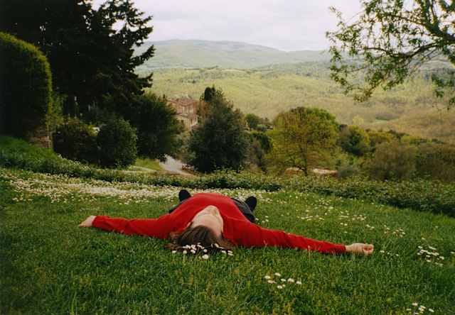 A man in a red jumper lies like a starfish on a hill, feet pointing to the wide Tuscan views, seen from grass above his head.
