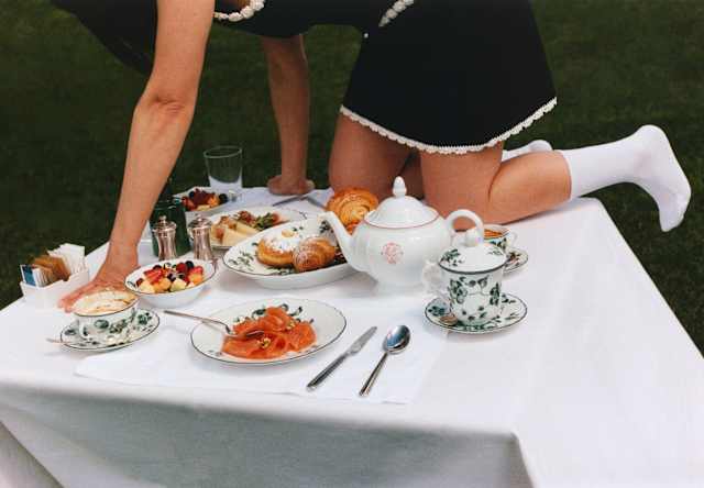 A woman in a black dress with white piping and socks crawls across a breakfast table with pastries, fruit, coffee and salmon.