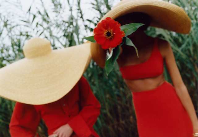Two women in red, one in a dress, the other in skirt and crop top, wear huge straw hats, obscuring their faces in the garden.