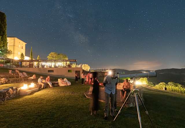 People gather on a grassy lawn at night, some near a firepit and others using a telescope, under a clear star-filled sky with visible buildings and distant hills.