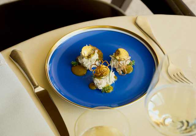 An Oro dish of three mounds of white rice, topped with edible gold discs and served with sauce on a gilt-rimmed blue plate.