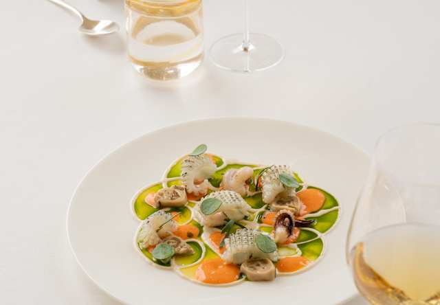 A white plate of pasta with green and orange vegetables sits on a round white table, accompanied by a glass of white wine, a glass of water, and cutlery, with a brown cushioned chair in the background.
