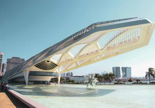 Futuristic white building with a geometric roof and open sides, set beside a shallow pool with a star-shaped sculpture; city skyline and palm trees in the background under a clear sky.