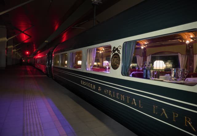 Lights glow in the restaurant car as the train waits on a platform with red and purple lights, seen at night from outside.