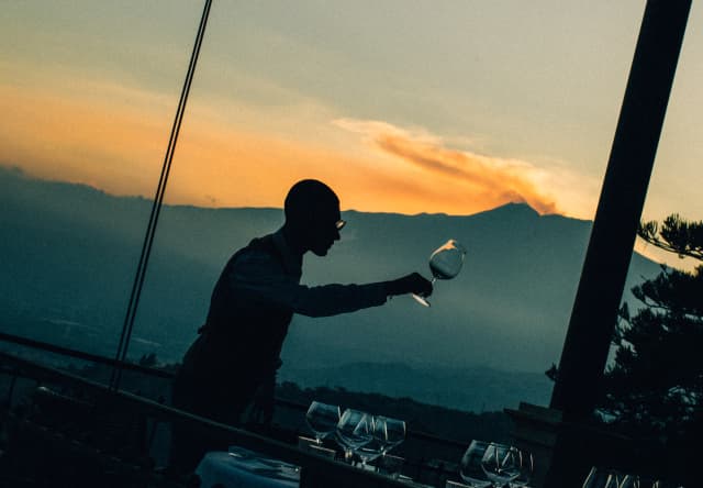 Angled image of a man selecting a wineglass from a table, barely visible in dusk light, with a sunset over Mount Etna behind.